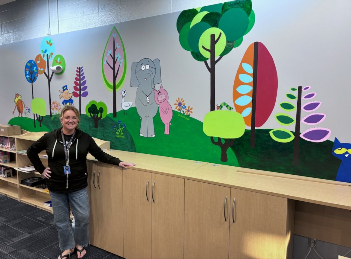 Mural with cute animal characters including a dog, elephant and pig, with Heidi Hansen standing in front of it